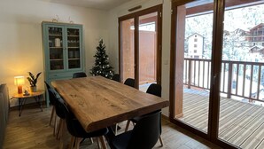 Un comedor con una mesa y sillas de madera, un aparador con puertas de cristal, un pequeño árbol de Navidad y un balcón con vistas a edificios y árboles.