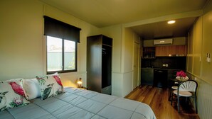 Ein Schlafzimmer mit Bett, Nachttisch, Fenster mit Jalousien, Kleiderschrank, einer Küchenzeile und einem Essbereich mit Stühlen.