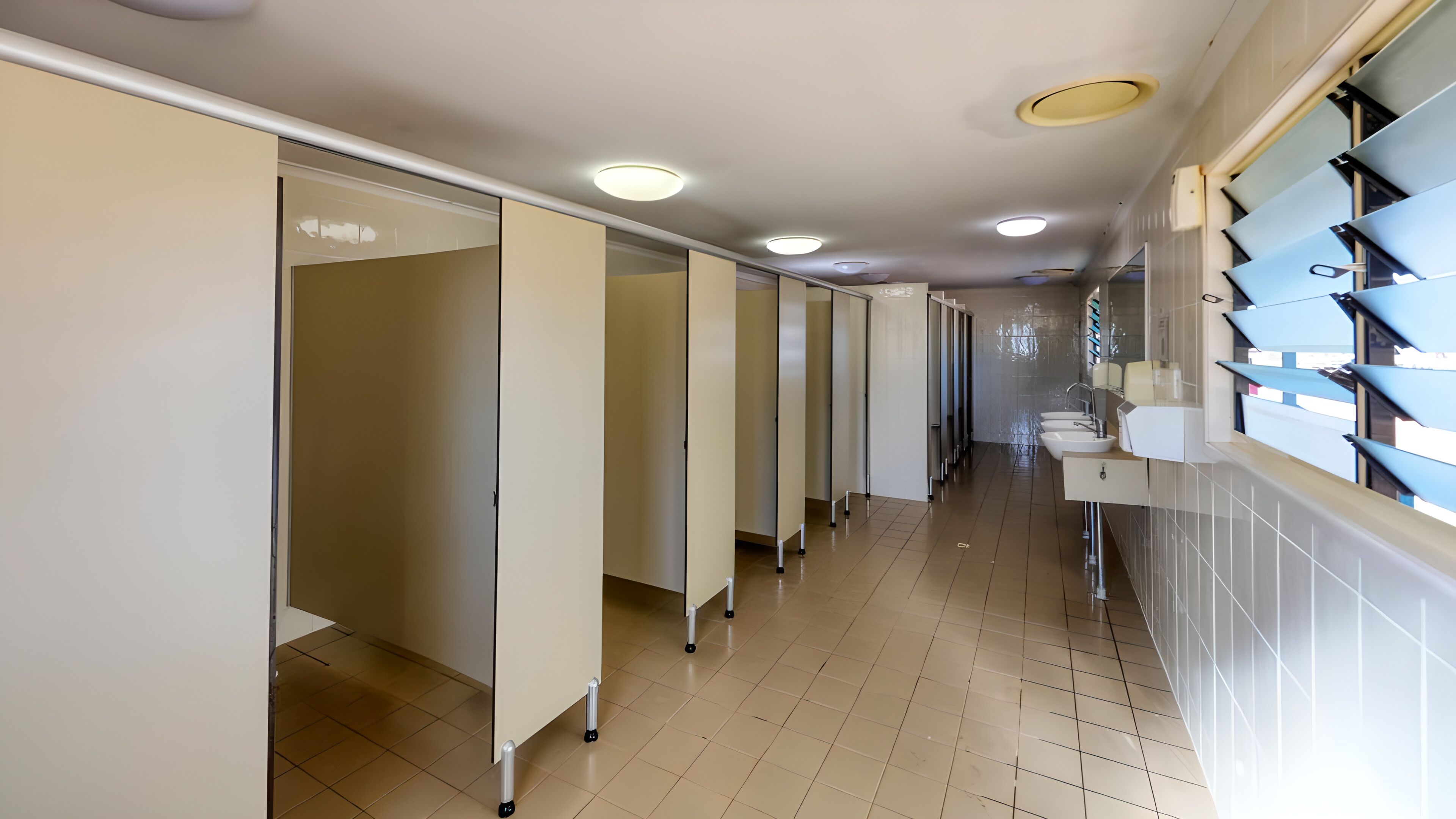 A hotel corridor with multiple doors, a tiled floor, and a row of sinks on the right.