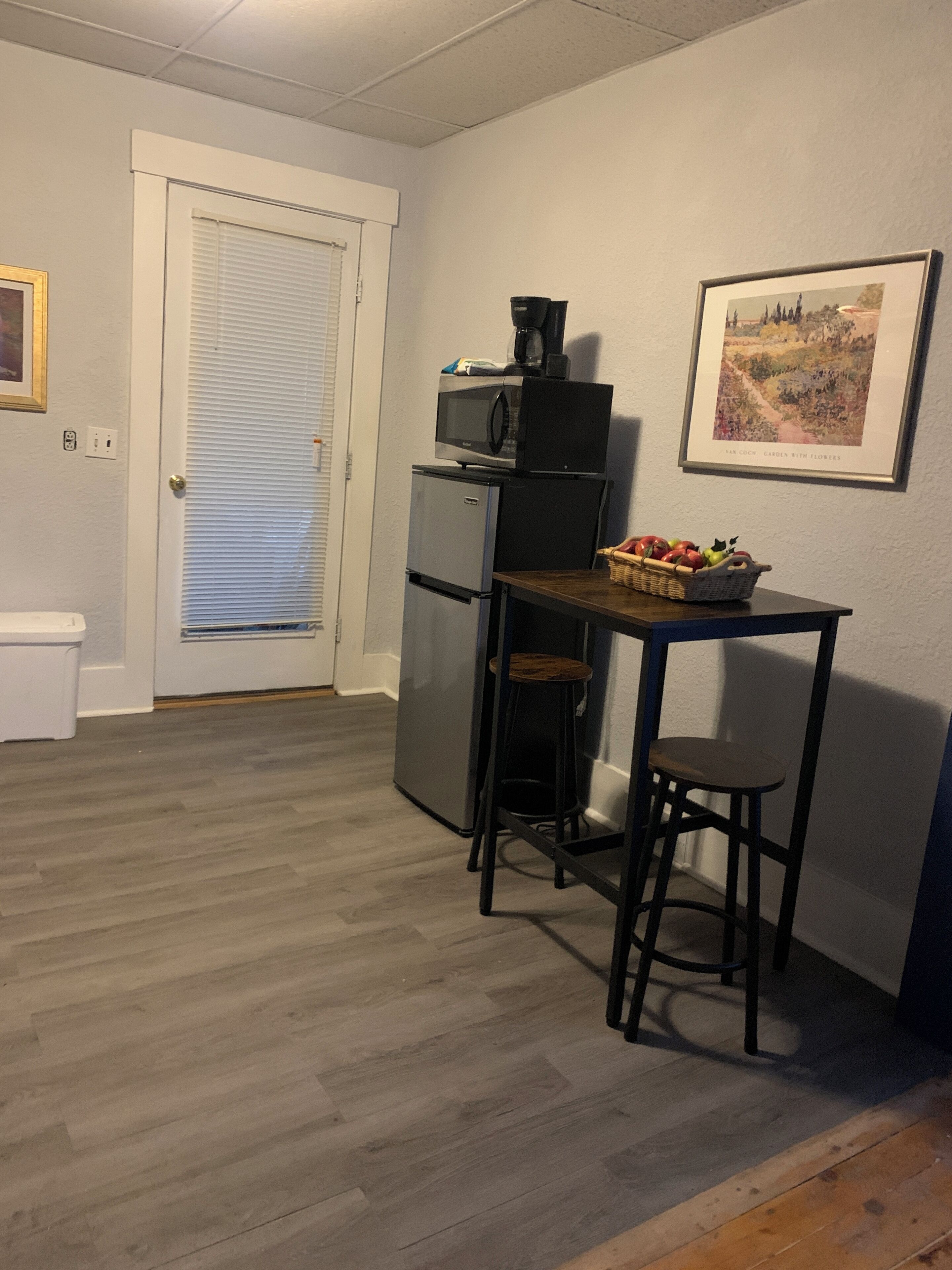 Een kleine keuken met een koelkast, magnetron en een eettafeltje met twee krukken. Er staat een mand met fruit op tafel, en aan de muur hangt een lijst.