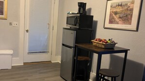 A compact kitchen with a refrigerator, microwave, and a small dining table with two stools. A basket of fruit is on the table, and a framed picture hangs on the wall.
