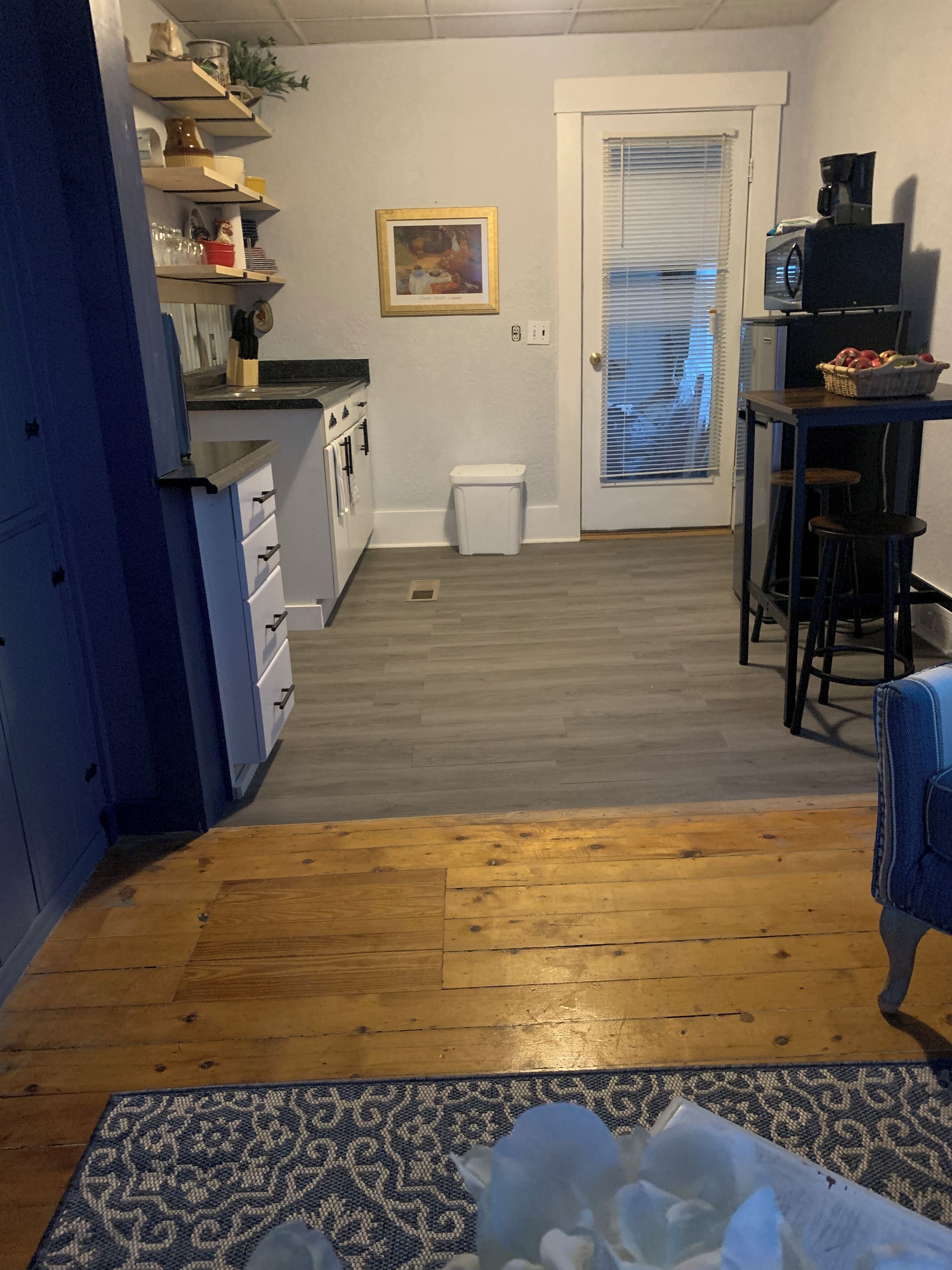 Een keuken met witte kastjes, een gootsteen en een aanrecht. Een prullenbak en een deur met raam zijn aanwezig.