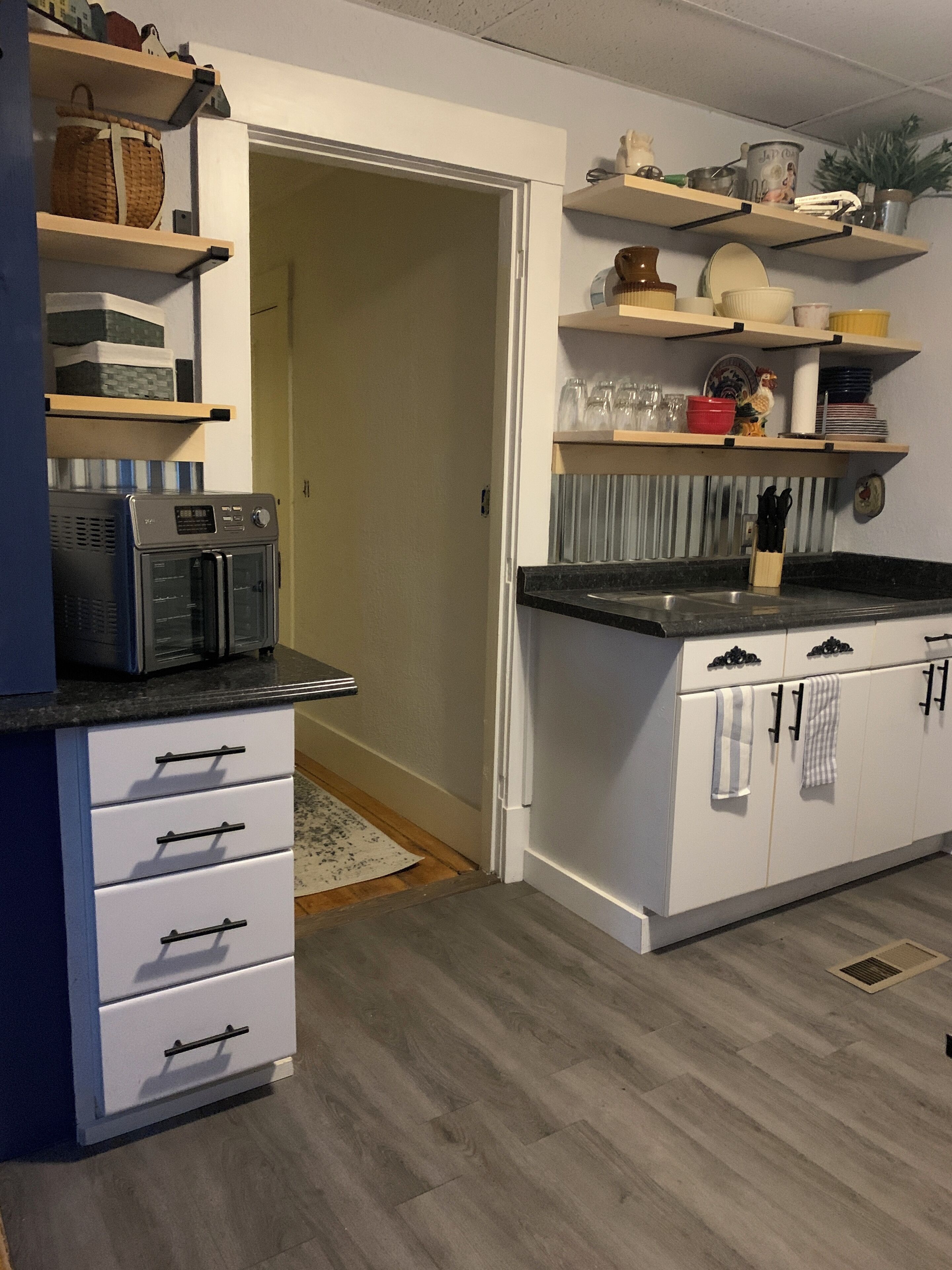 Een keuken met witte kasten, een zwart aanrecht en houten vloer. Boven het aanrecht zijn planken met verschillende keukengerei.