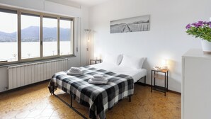 A bedroom with a bed, a window with a view of mountains, a bedside table with a lamp, and a wall-mounted picture. - Lago 2 - Three Bedroom Apartment, Sleeps 8 (Oggebbio)