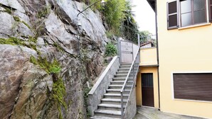 A staircase leading up a rocky hillside, adjacent to a yellow building with a closed shuttered door. - Lago 2 - Three Bedroom Apartment, Sleeps 8 (Oggebbio)