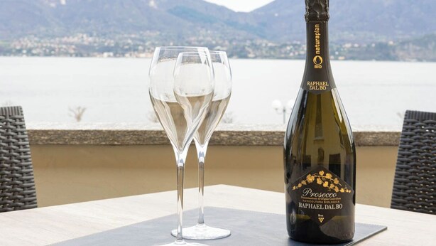 A bottle of Prosecco on a table with two empty wine glasses, set against a scenic mountain backdrop. - Lago 2 - Three Bedroom Apartment, Sleeps 8 (Oggebbio)