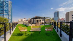 Eine Dachterrasse mit grünem Kunstgrashalm, Korbstühlen und einem überdachten Sitzbereich.