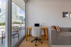 Une pièce moderne avec une grande fenêtre, un bureau, un fauteuil de bureau gris, un canapé gris et deux tableaux encadrés accrochés au mur.