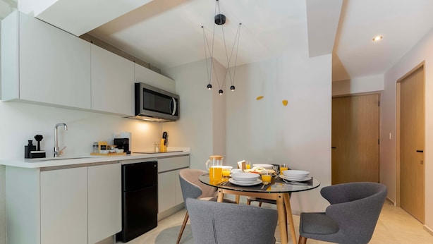 Una cucina moderna con mobili bianchi, una lavastoviglie nera e un forno a microonde. C'è un tavolo da pranzo con due sedie grigie e un piano in vetro. Un lampadario pende dal soffitto, e si intravede una porta in legno sullo sfondo.