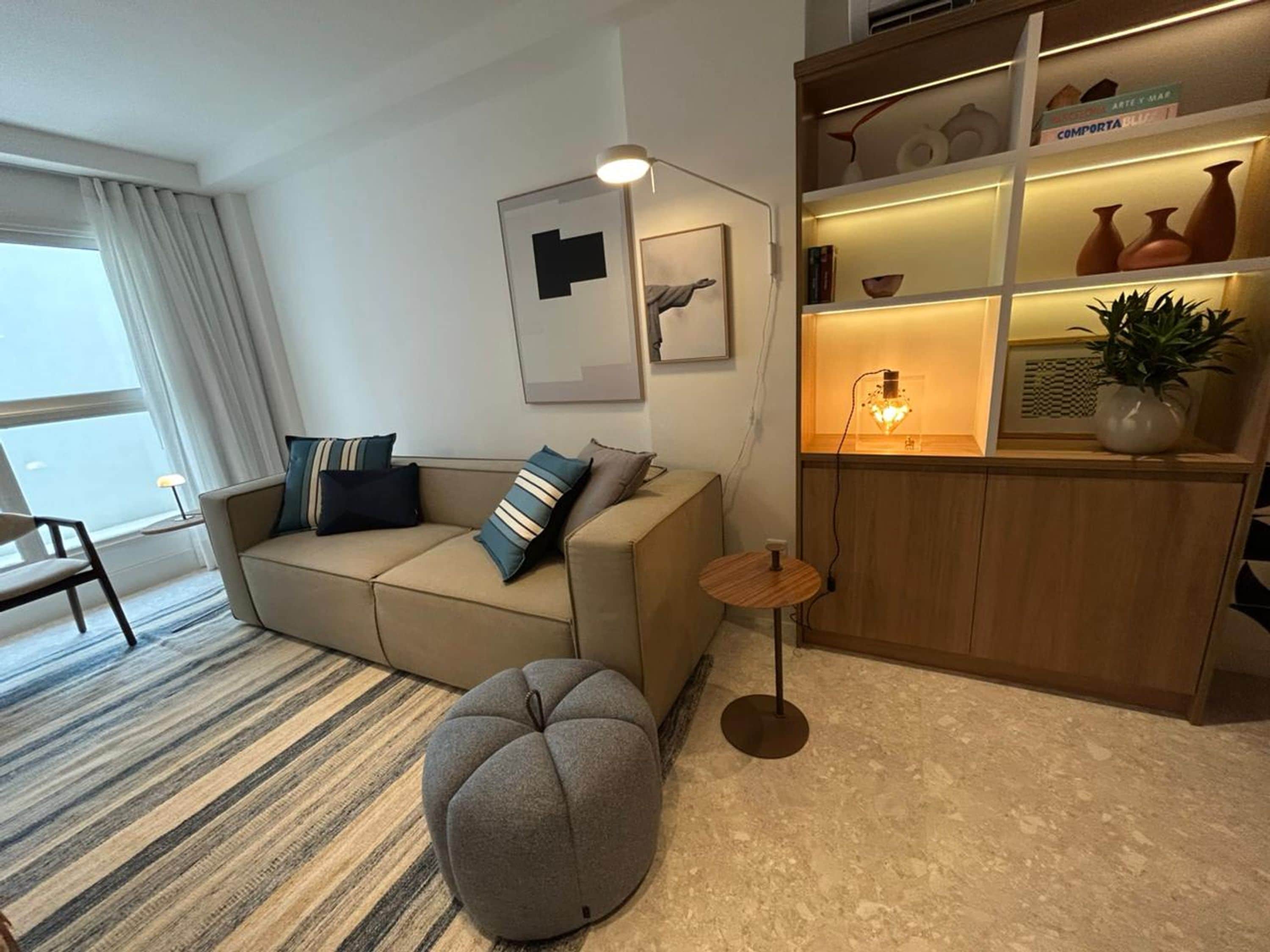 Ein modernes Wohnzimmer mit einer Couch, einem Hocker und einem Beistelltisch. Ein Bücherregal ist mit Dekoration und einer Lampe bestückt.