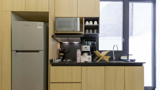 Eine kleine Küche mit Kühlschrank, Mikrowelle, Kaffeemaschine und einem Fenster mit Tageslicht.
