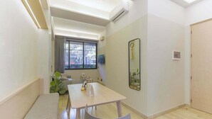 Ein kleiner, gut beleuchteter Raum mit Holzboden, einem weißen Tisch und einem einzelnen weißen Stuhl. Es gibt ein Fenster mit Vorhängen, einen kleinen Spiegel an der Wand und eine Tür auf der rechten Seite.