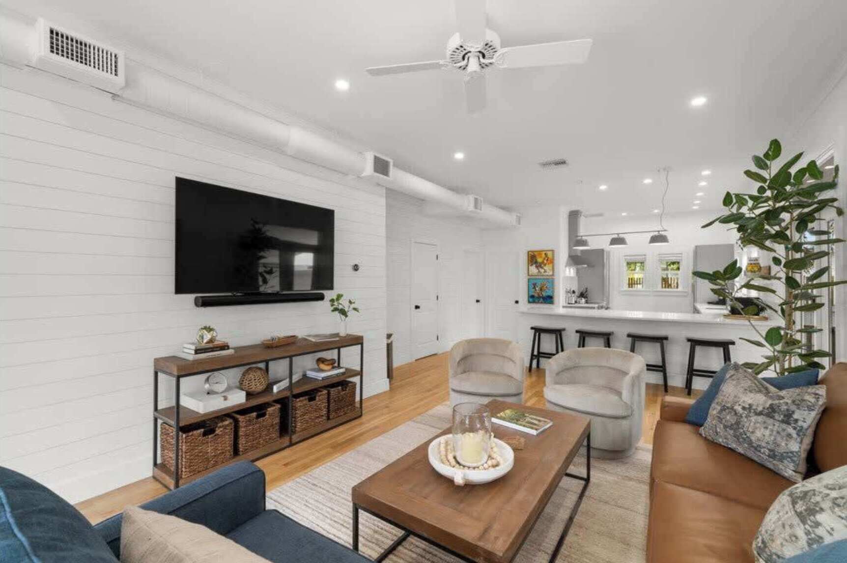 A modern living room with a flat-screen TV, comfortable seating, a coffee table, and a kitchen area in the background.