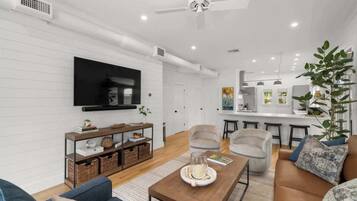 A modern living room with a flat-screen TV, comfortable seating, a coffee table, and a kitchen area in the background.