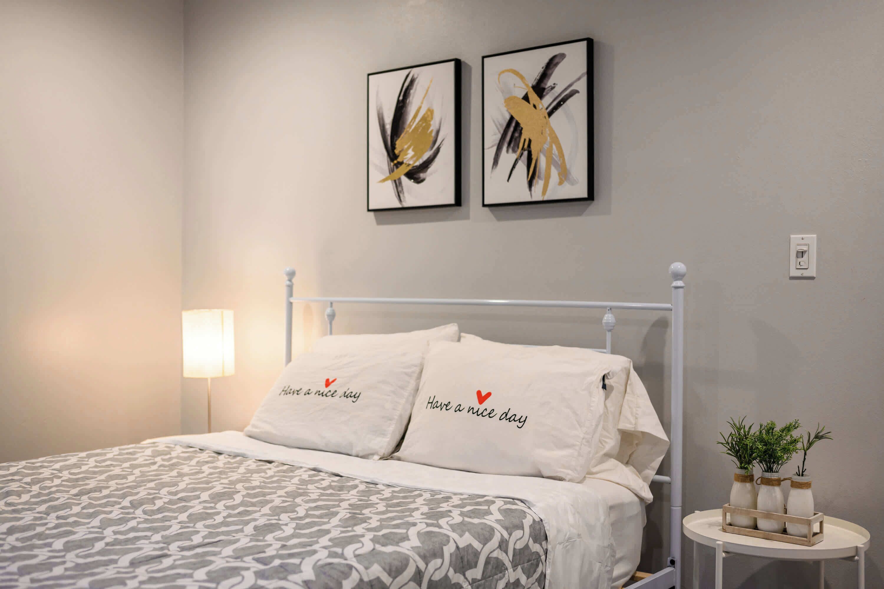 A neatly made bed with patterned bedding and two pillows with heart messages, a bedside table with decorative vases, and abstract art on the wall.