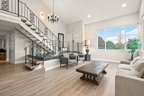 Amplia sala de estar con piso de madera, una escalera con barandas negras, una araña y un ventanal con vistas, además de un sofá color crema.