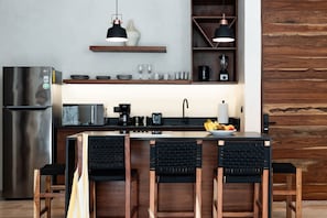 A modern kitchen with a stainless steel refrigerator, a coffee maker, and a sink. There are three wooden chairs with black cushions and a wooden island with a bowl of fruit. - Bohemian 2BR Boutique Condo in Trendy La Veleta (Tulum)