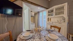 A dining area with a table set for four, a television mounted on the wall, and a kitchen area with cabinets and a window. - Small country cottage with wifi and parking (Auguaise)