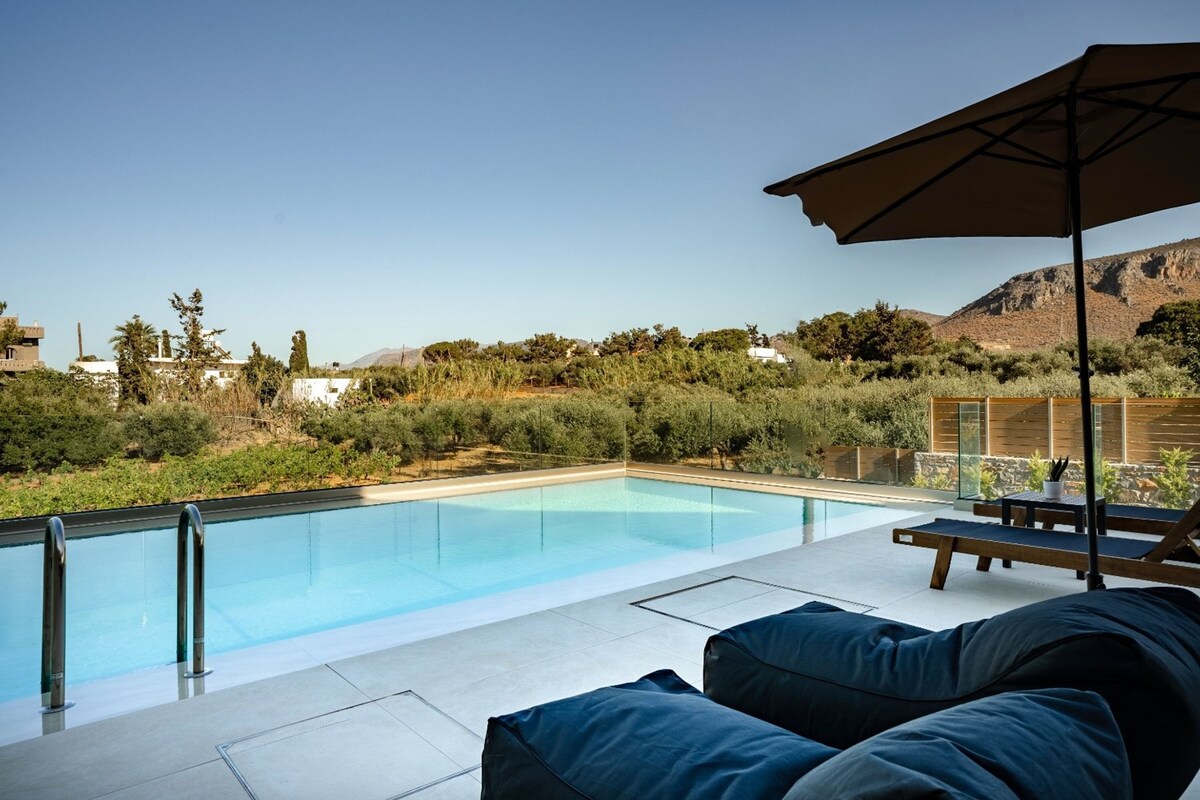 A modern outdoor pool area with clear blue water, surrounded by lush greenery and a mountain backdrop.