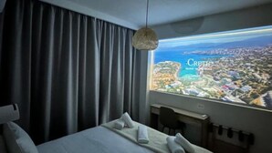 A hotel room with a bed, a desk, and a view of a coastal city. - Nerites Luxury Villas (Hersonissos)