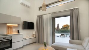 A modern living room with a sofa, a coffee table, and a TV. There is a kitchen area with cabinets and a built-in oven. - Nerites Luxury Villas (Hersonissos)