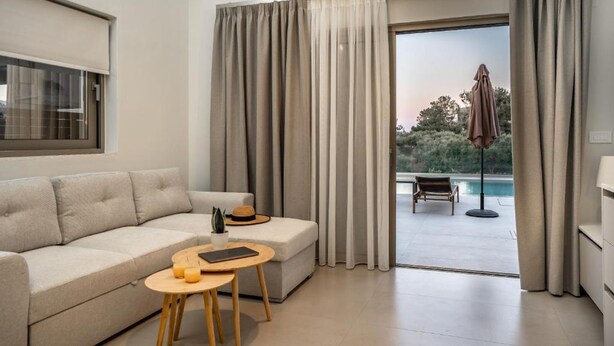 A modern living room with a sofa, a small table, and a view of an outdoor pool area. - Nerites Luxury Villas (Hersonissos)