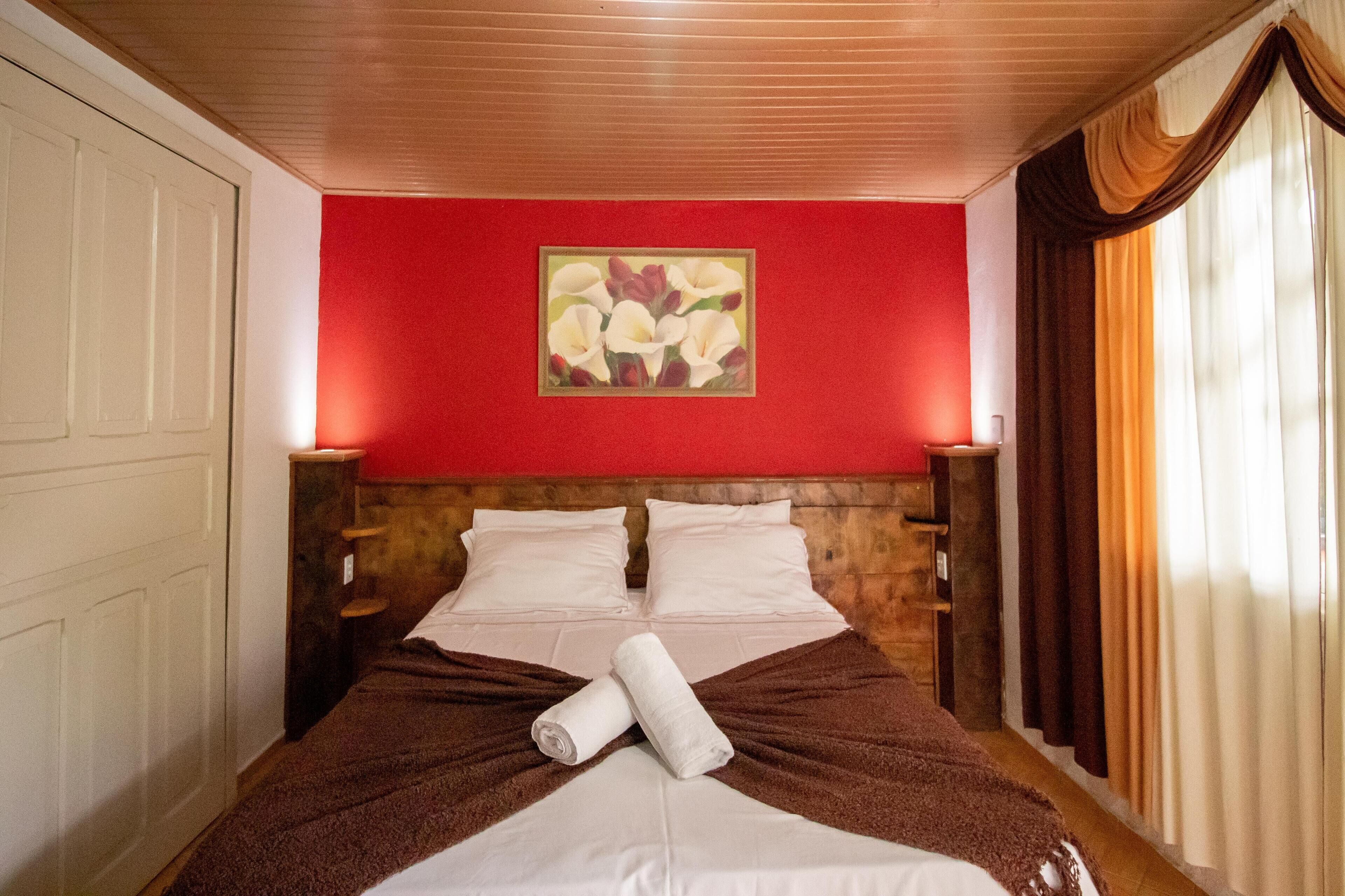 Quarto de hotel com cabeceira de madeira, parede em vermelho e um quadro sobre a cama.