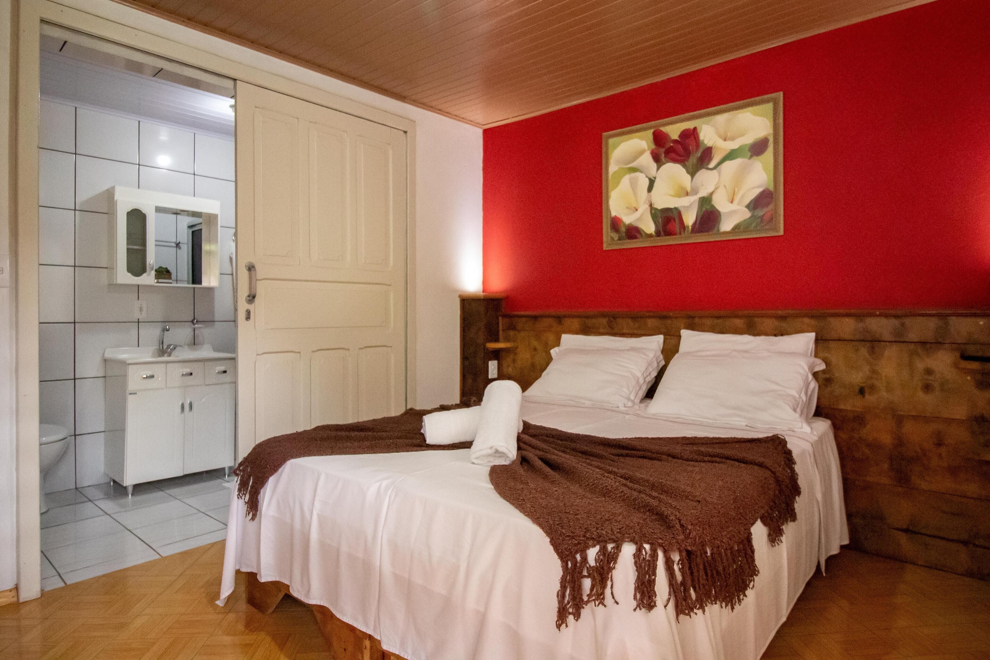 A bedroom with a wooden headboard, a white bed with a brown blanket, a red wall with a floral painting, and a white bathroom with a mirror and cabinets.