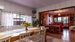 A dining area with a wooden table and white chairs, a vase of flowers, and a view of the outdoors through large windows. - Hospedagem Rural Recanto do Riacho (Urubici)
