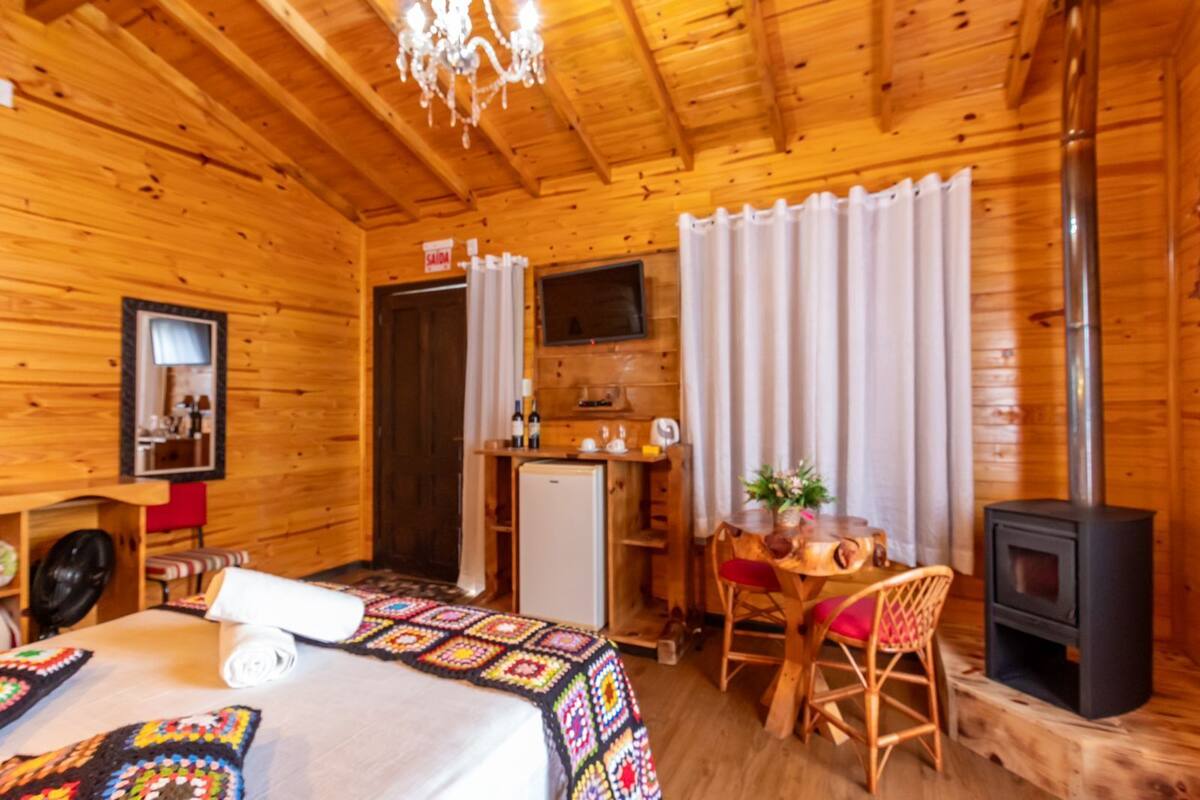 A cozy wooden cabin room with a bed, a desk, a chair, a small refrigerator, a television, and a wood-burning stove.