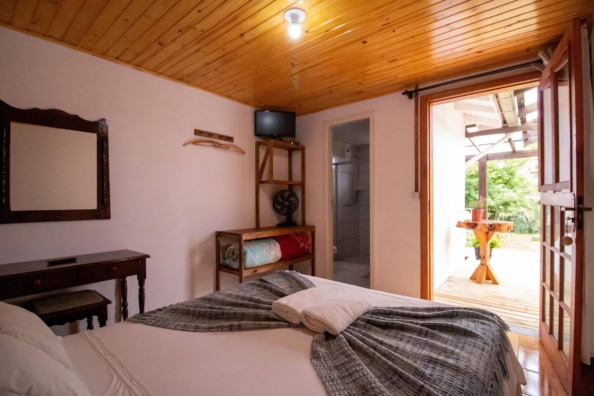 A bedroom with a wooden ceiling, a bed with a blanket, a bedside table, a TV on a stand, and a view of the outside through a glass door.