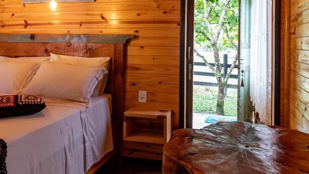 A wooden cabin room with a bed, a table, and a door leading outside. - Hospedagem Rural Recanto do Riacho (Urubici)