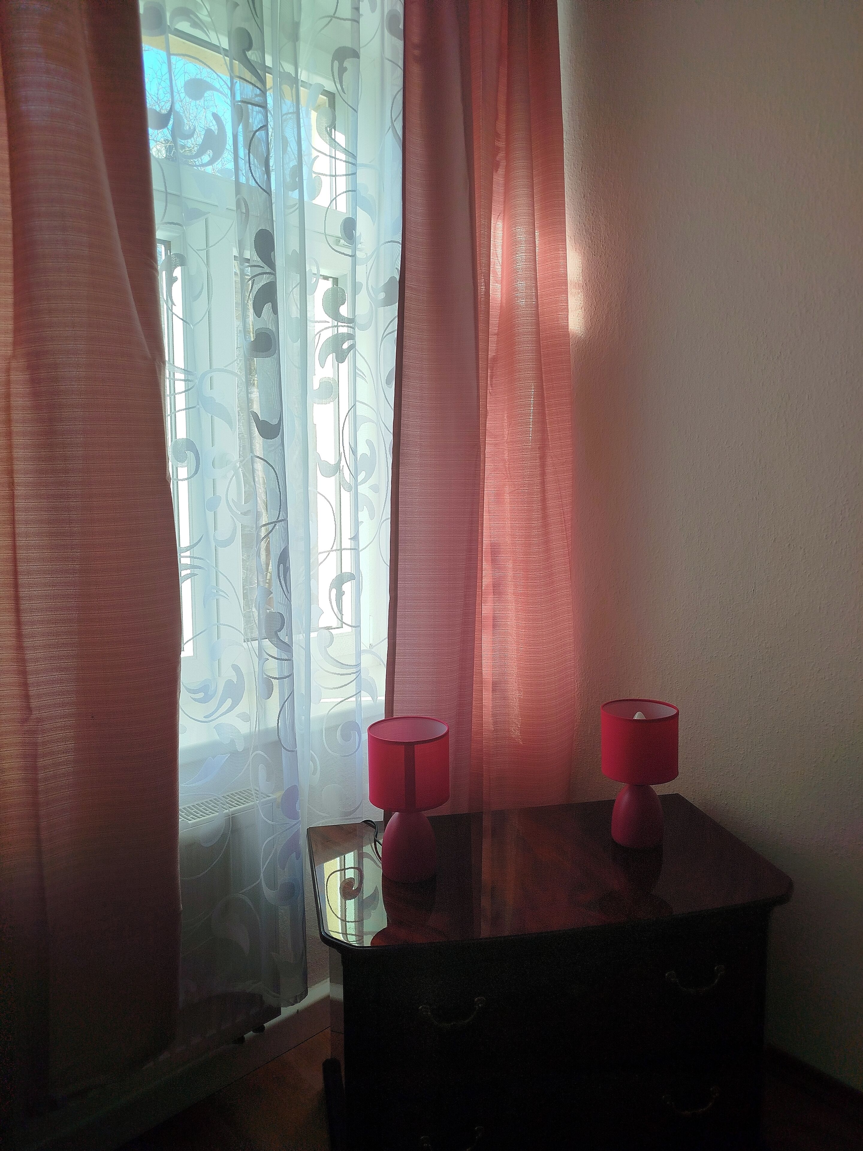 Una habitación con una ventana cubierta por cortinas de gasa y un cortinaje estampado. Hay una mesita con una lámpara roja encima.