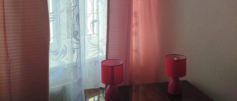A room with a window covered by sheer curtains and a patterned curtain. There is a small table with a red lamp on it.