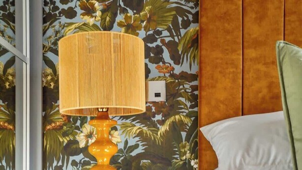 Habitación de hotel con papel tapiz floral, cabecera de madera, mesita de noche con lámpara y cama con almohadas.