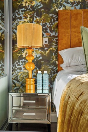 Quarto de hotel com papel de parede floral, cabeceira de madeira, mesa de cabeceira com abajur e cama com almofadas.