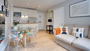 Een moderne woonkamer met een glazen eettafel, transparante stoelen, een witte bank met geometrische kussens, en een keuken op de achtergrond.