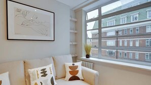 Een moderne woonkamer met een beige bank, abstracte kussens, een ingelijste kunstwerk en uitzicht op een bakstenen gebouw door een groot raam.