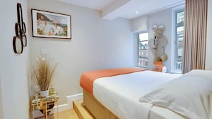 Een moderne slaapkamer met een bed, een nachtkastje met een vaas, een raam met gordijnen en uitzicht op een stadsbeeld.