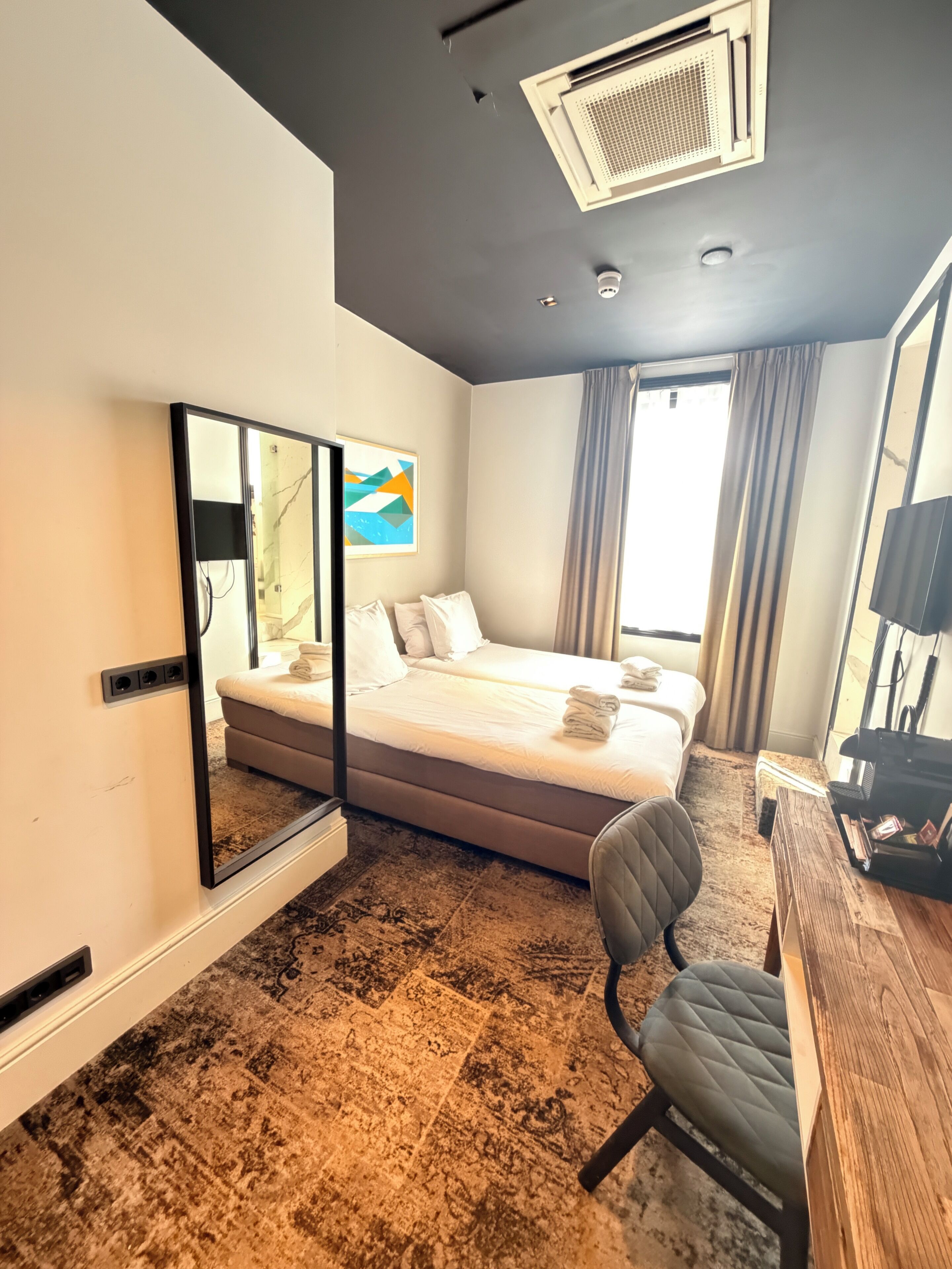 A hotel room with a bed, a desk, a chair, a large mirror, a ceiling fan, and a window with curtains.