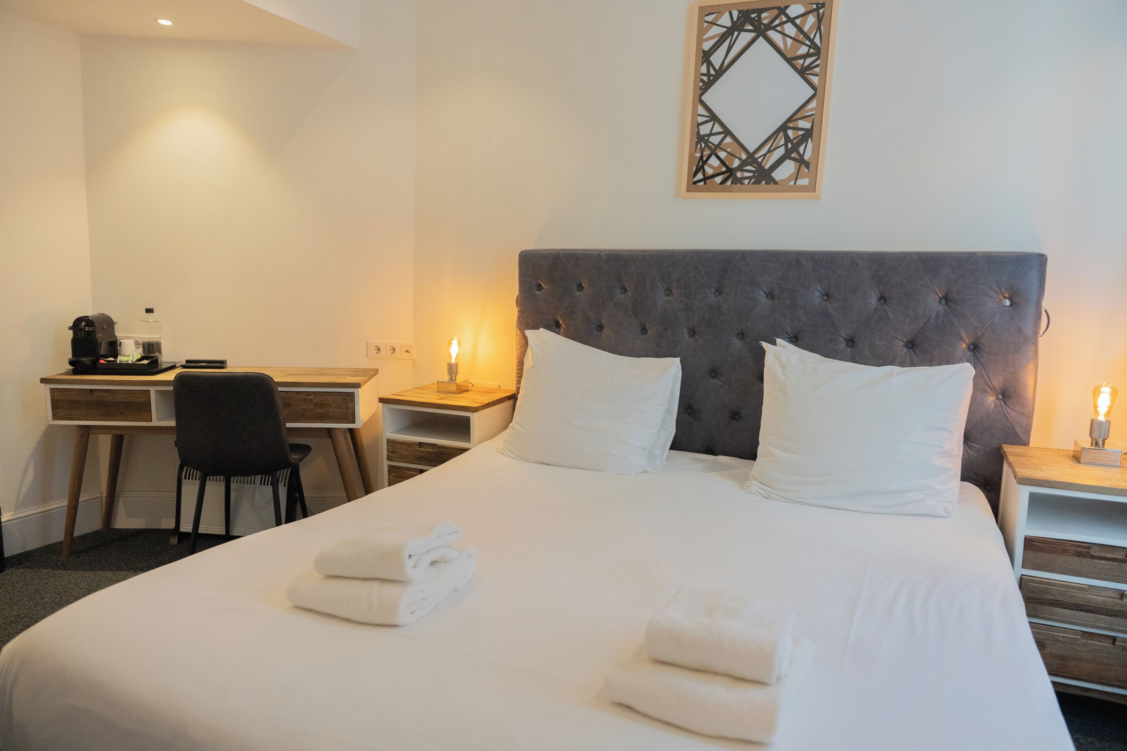 Une chambre d’hôtel comprenant un lit, deux tables de chevet avec des lampes, un bureau avec une chaise et un élément décoratif mural.