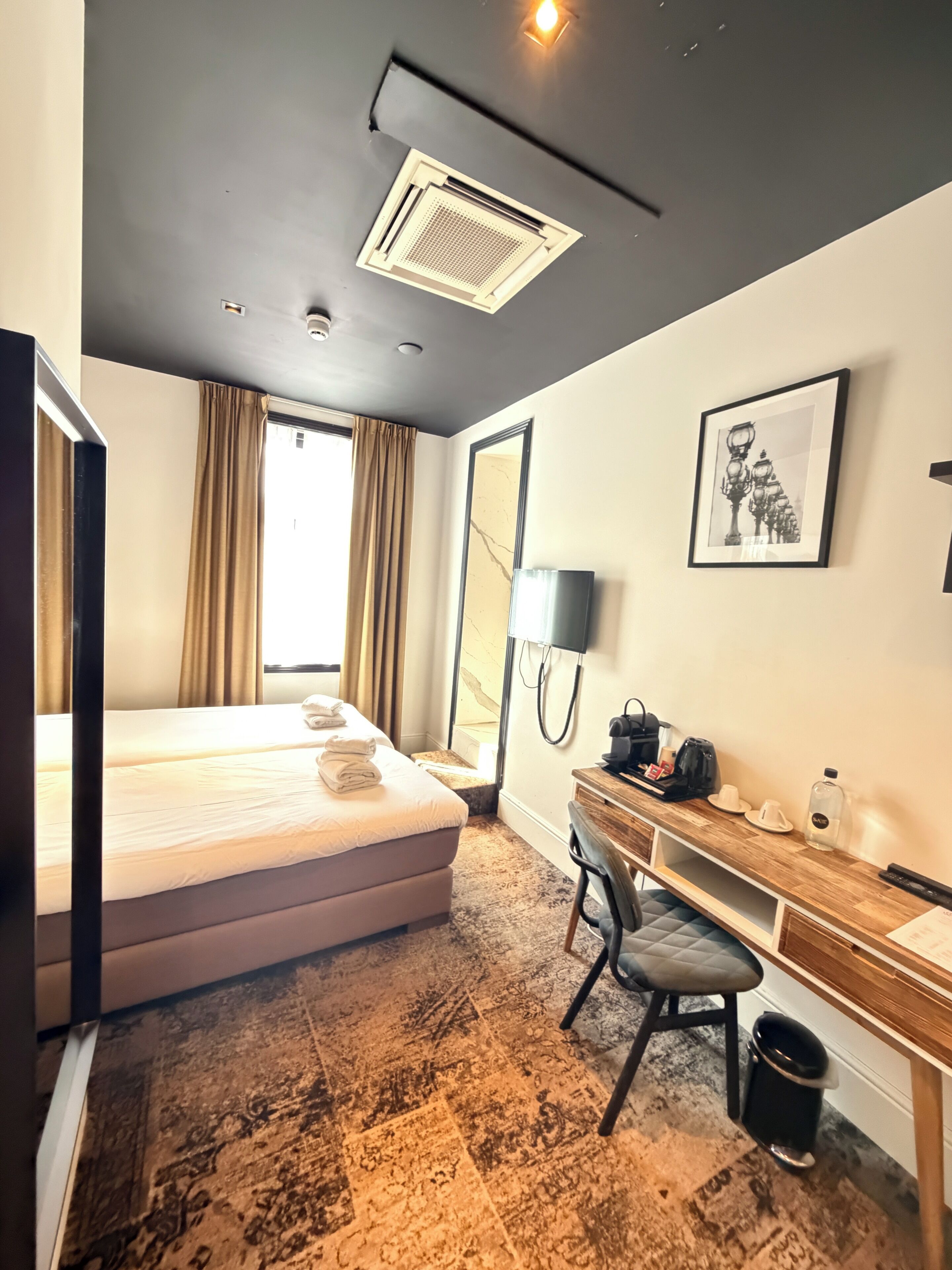 A hotel room with a bed, a desk, a chair, a trash bin, a lamp, and a framed picture on the wall.