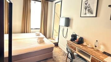 A hotel room with a bed, a desk, a chair, a trash bin, a lamp, and a framed picture on the wall.