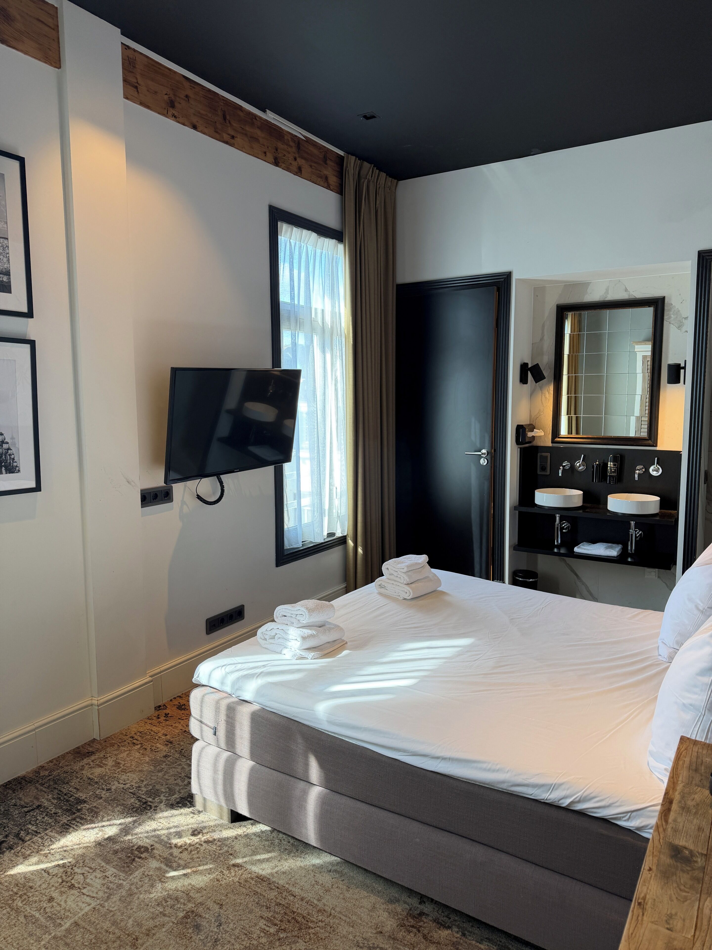 A hotel room with a bed, a TV mounted on the wall, a bathroom with a mirror, and a window with curtains.