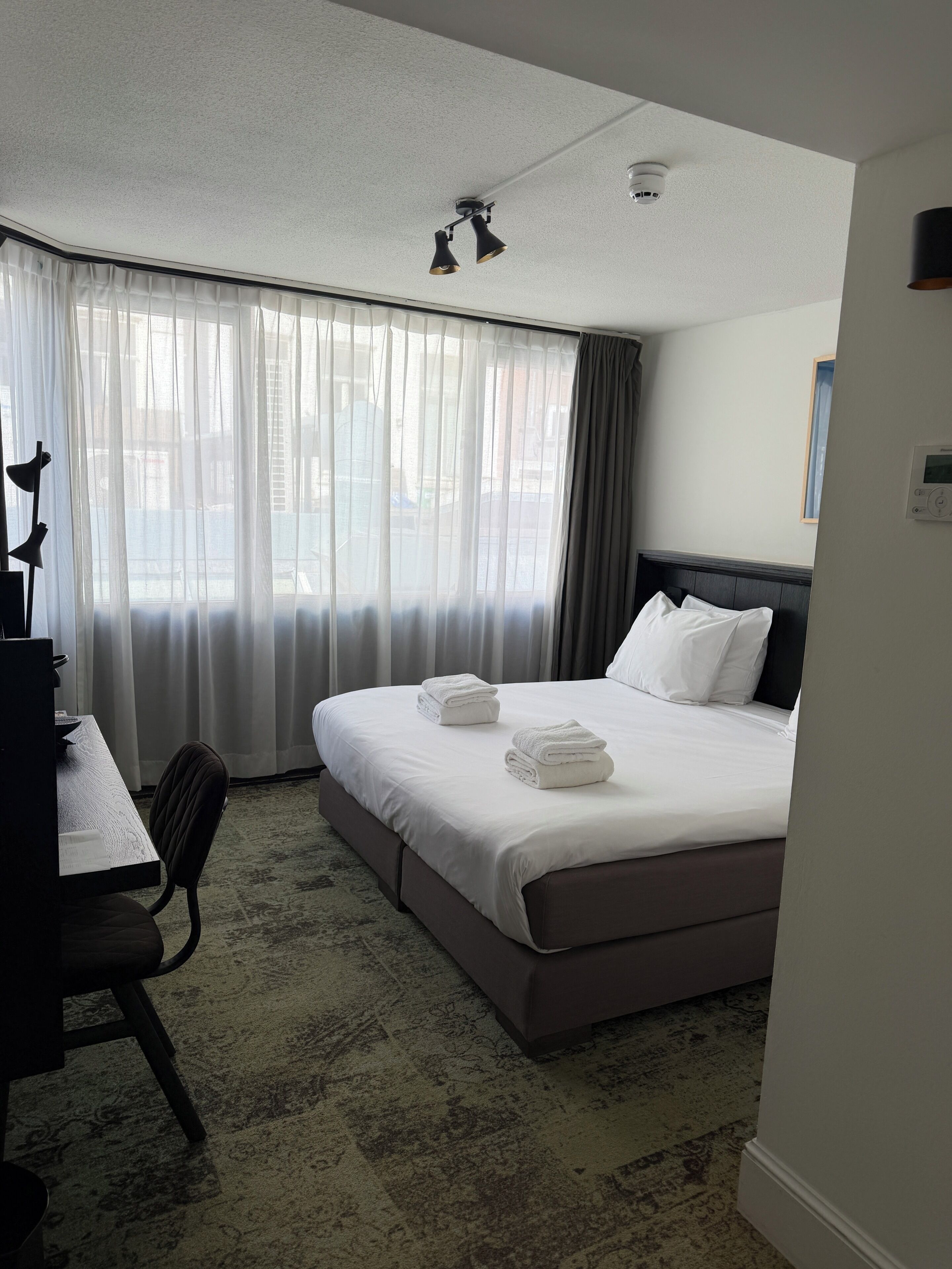 A hotel room with a bed, a desk, a chair, a window with curtains, and a ceiling light fixture.