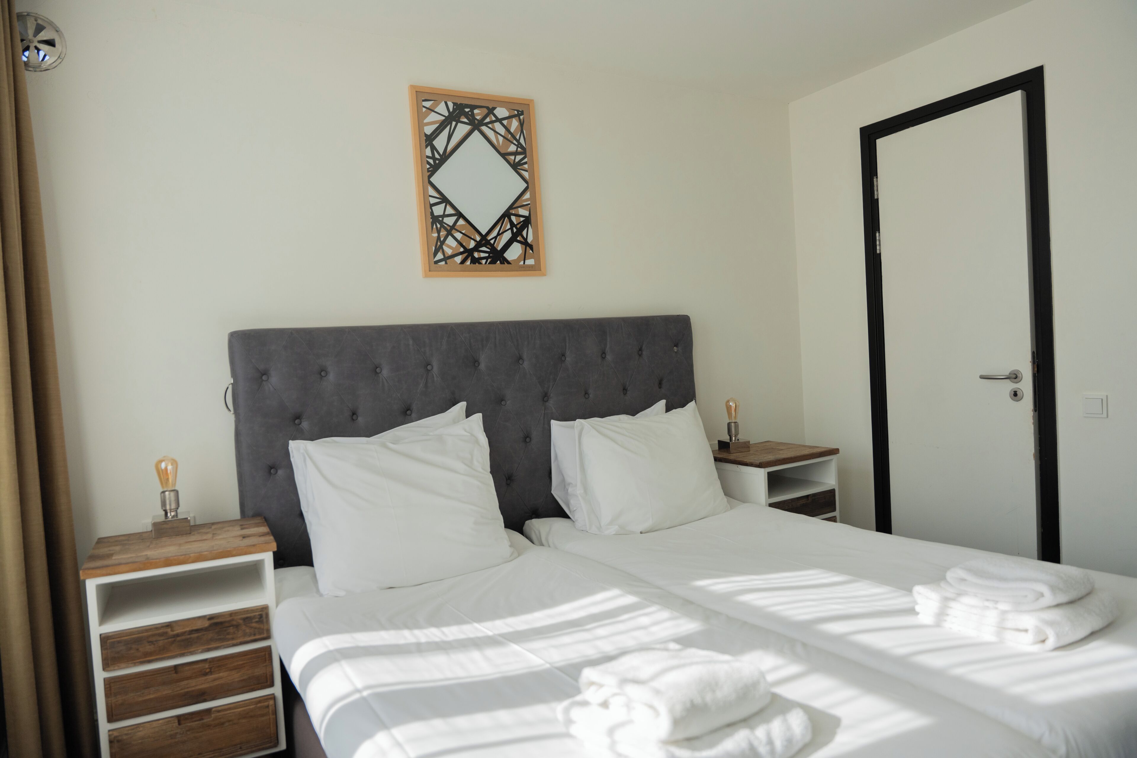 A neatly made bed with white linens, a grey upholstered headboard, a wooden nightstand with a lamp, and a wall-mounted decorative mirror.