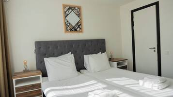 A neatly made bed with white linens, a grey upholstered headboard, a wooden nightstand with a lamp, and a wall-mounted decorative mirror.