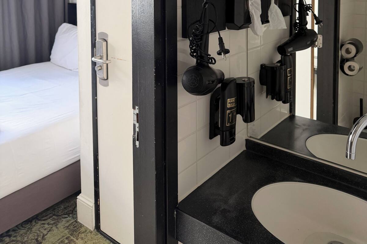 Une chambre d’hôtel comprenant un lit, une salle de bain avec un lavabo et un miroir, ainsi qu’un sèche-mains.