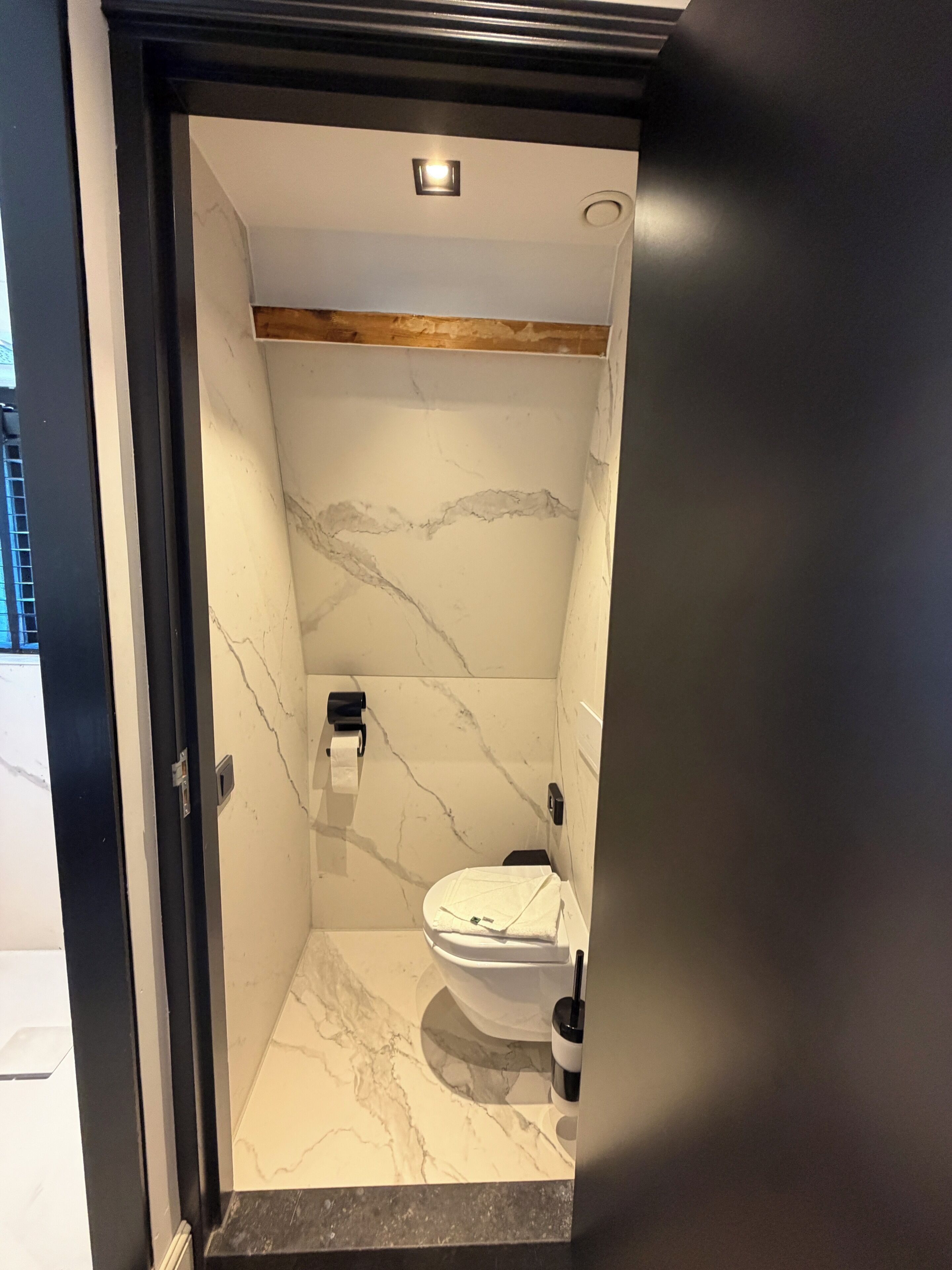 Une salle de bain avec un mur en marbre, des toilettes, un distributeur de serviettes et un plafond en bois.