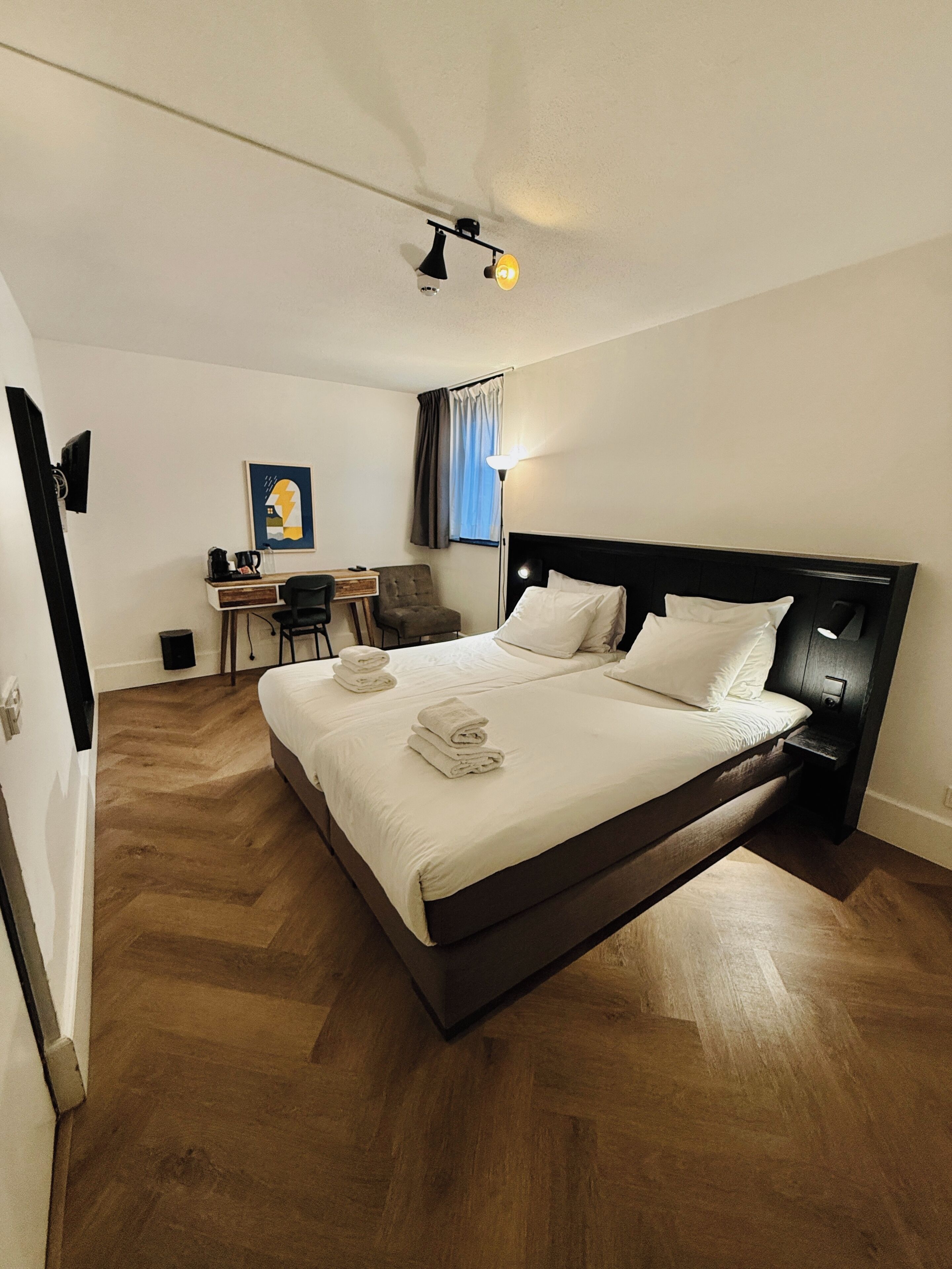 A hotel room with a bed, bedside table, chair, desk, and a painting on the wall.